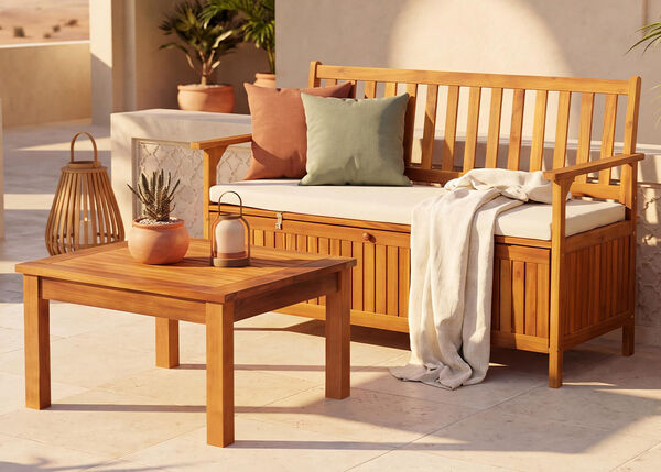 Cozy outdoor patio with a wooden storage bench, coffee table, cushions, potted plants and warm natural decor.