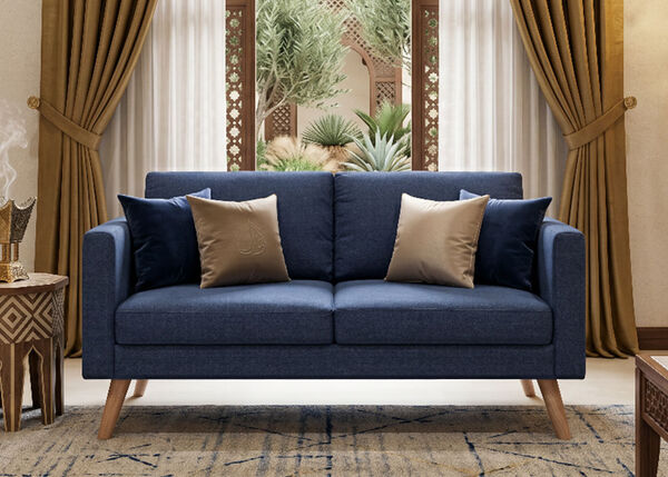 A navy blue sofa with three decorative pillows sits in a stylized room setting featuring sheer curtains and greenery. The pillows are tan/gold with an embossed design and dark blue velvet.