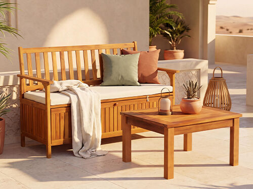 Cozy outdoor patio with a wooden storage bench, coffee table, cushions, potted plants and warm natural decor.