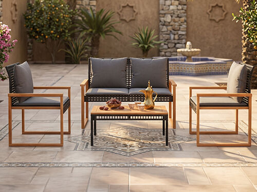 Contemporary outdoor furniture set - a sofa, two armchairs, and a coffee table &ndash; arranged in a traditional Arabic courtyard with a decorative coffee pot and dates. The setting includes a stone wall with patterned details and lush greenery. Click to see the products in the collection.