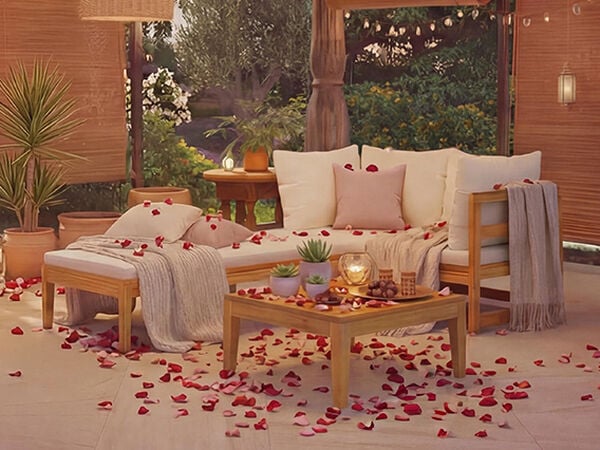Outdoor seating area with a wooden sofa, pillows, blankets, and a small table, decorated with rose petals scattered on the floor. Click to see the products in the collection.