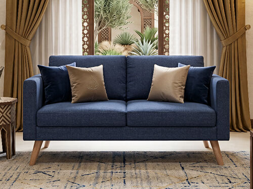 A navy blue sofa with three decorative pillows sits in a stylized room setting featuring sheer curtains and greenery. The pillows are tan/gold with an embossed design and dark blue velvet. Click to see the products in the collection.