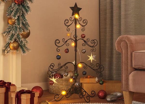 Black metal Christmas tree decorated with baubles, lights and stars, with gift boxes and ornaments on the floor. An armchair and a fireplace with decorated garlands in a styled living room. 