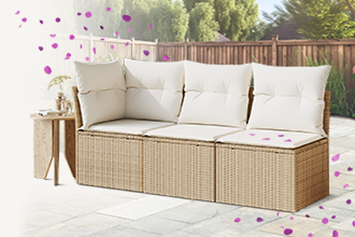 In a courtyard, a light brown woven rattan modular sofa with a matching side table, cream-colored cushions and a throw, with purple confetti. Click to discover more products.