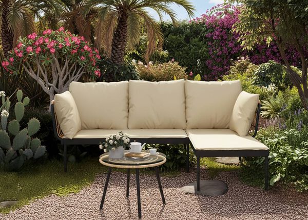 A black outdoor lounge set with cream cushions, a glass-top side table, in a lush garden with palm trees, cacti, and blooming pink flowers.