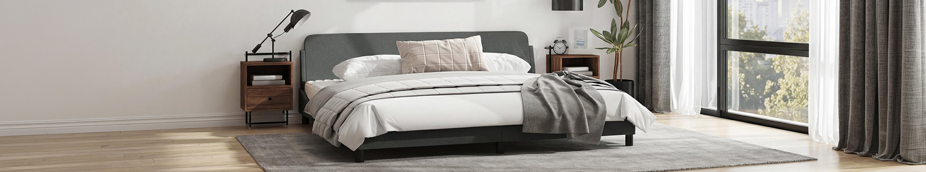 A modern bedroom interior featuring a dark gray upholstered bed with white linens and a gray throw blanket. A small wooden nightstand with books resides on either side of the bed. Curtains and a large window flank the bed, overlooking a building. A gray area rug is centered beneath the bed..
